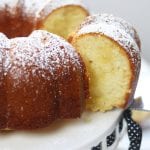 A slice being lifted from a Lemon Pound Cake on a white cake stand, wrapped with a black ribbon adorned with white polka dots.