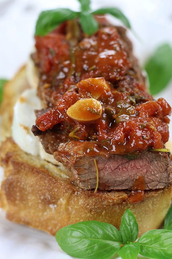 Steak Pizzaiola onto of garlic toast with mozzarella on a white plate.