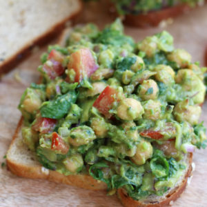 Up close photo of an open Avocado Chickpea Sandwich on a brown cutting board next to two slices of tomatoes.