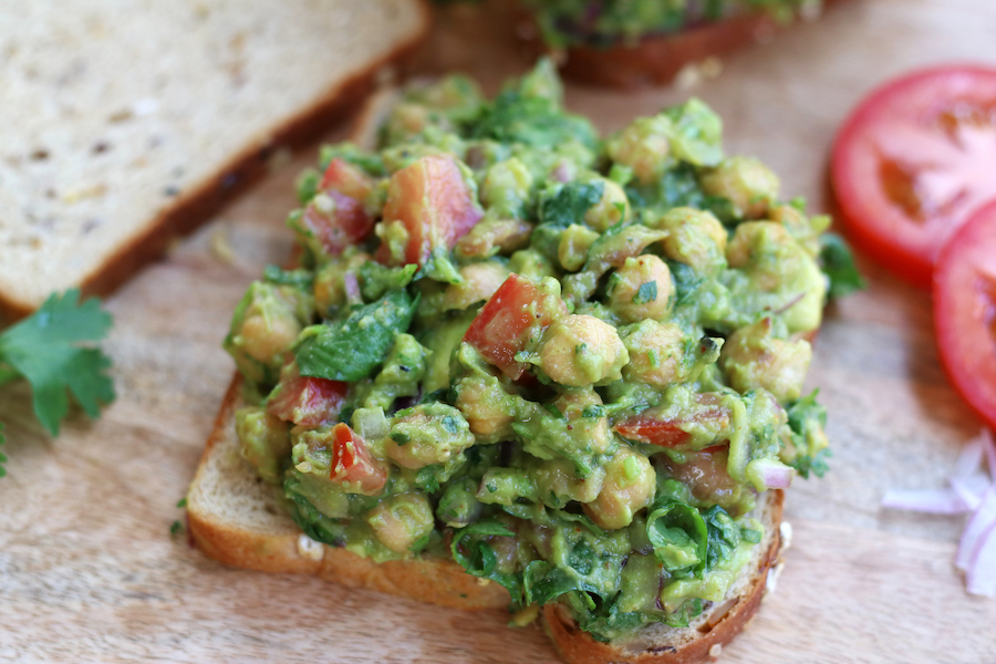 Up close photo of an open Avocado Chickpea Sandwich on a brown cutting board next to two slices of tomatoes.