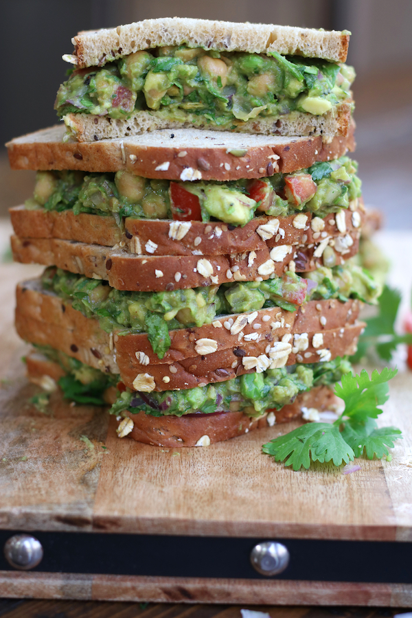 A stack of four Guacamole Sandwiches with seeded whole grain bread on a wooden cutting board.