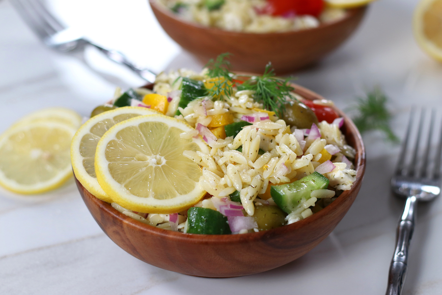 A small bowl of Orzo Pasta Salad garnished with two lemon slices and a fork sitting next to the bowl.