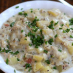 New England Clam Chowder in a white bowl sitting next to a soup spoon.