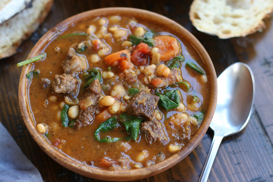 A serving of Bean and Beef Stew sitting on a wooden table with a spoon and bread slices.