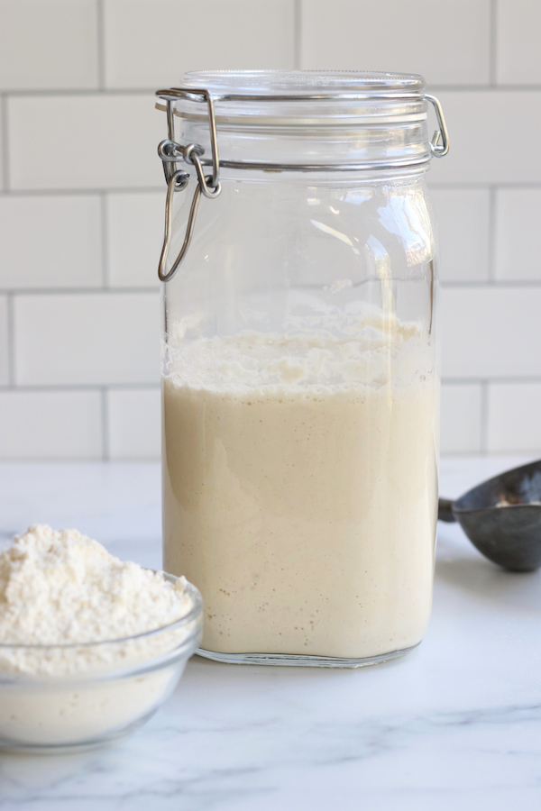 Sourdough Bread Starter in a glass jar sitting in a white countertop with a bowl of flour and a scoop.