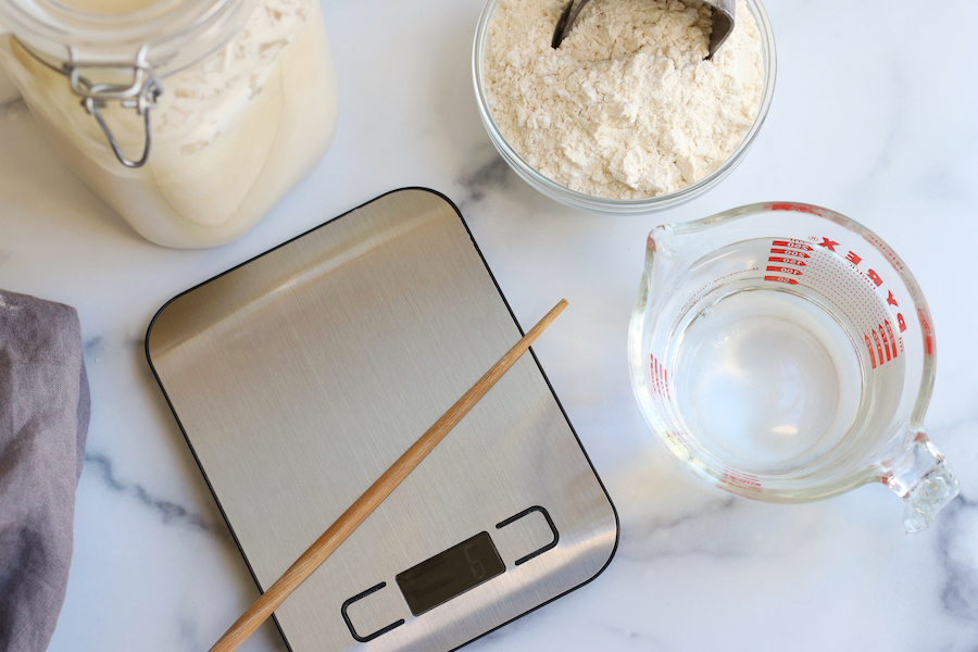 Overhead photo of baking scale, liquid measuring cup, chopstick and glass bowl; tools needed to make sourdough starter.