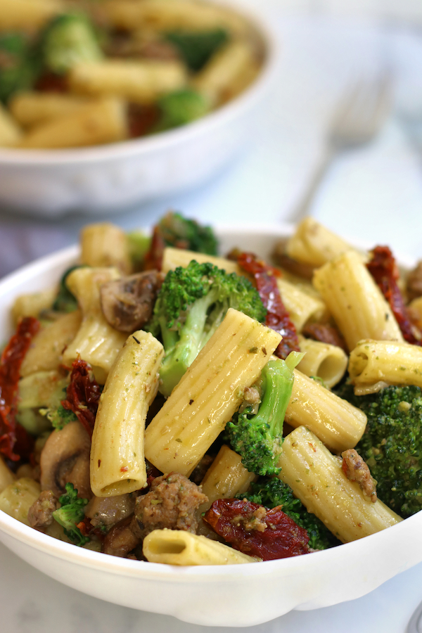 Two bowls of pasta with pesto, sausage, broccoli and sun-dried tomatoes.