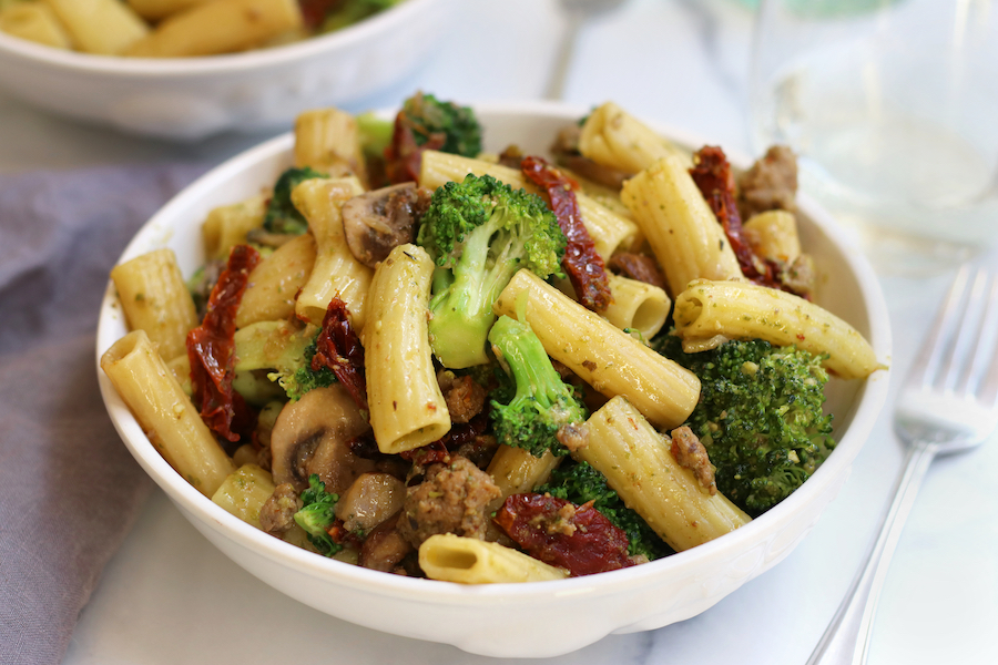 A serving of Pesto Pasta with Sausage sitting on a white countertop with a fork and glass of wine.