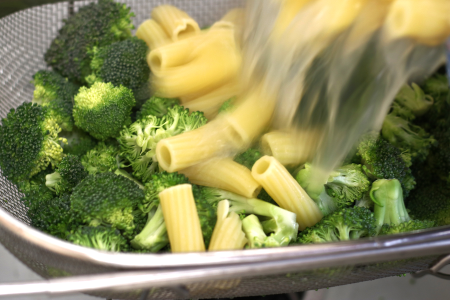 Pouring cooked pasta over broccoli florets for Sausage Pesto Pasta.