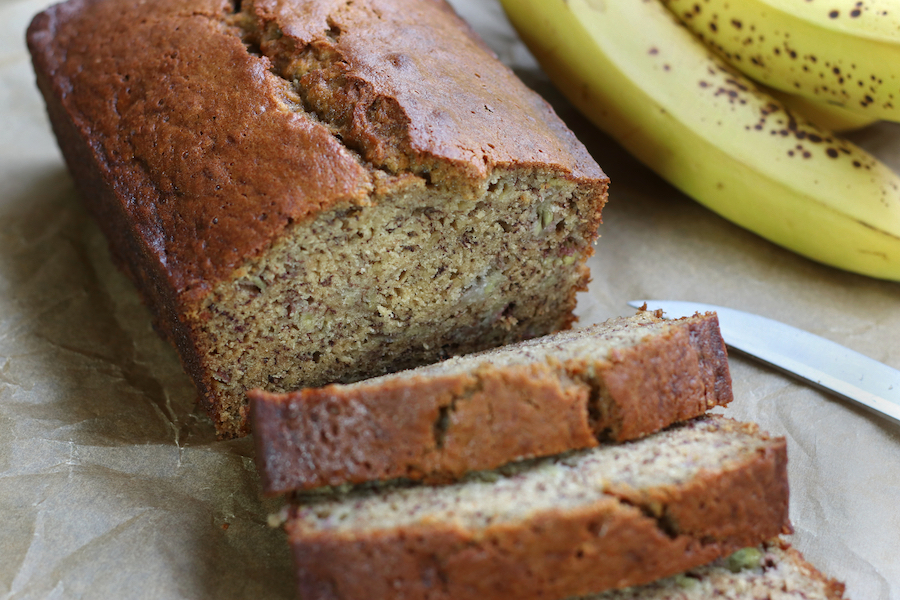 A loaf of sliced Banana Bread made with oil sitting on brown parchment paper next to ripe bananas.