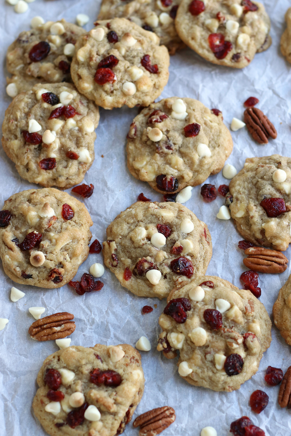 An over photo of Cranberry Cookies on white parchment paper with pecans, cranberries and white chocolate chips scattered around them.