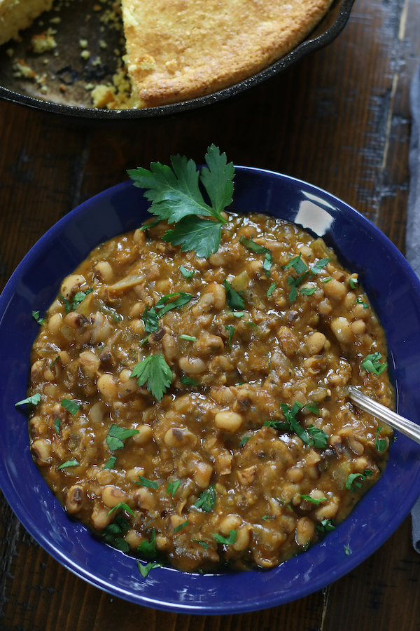 A blue bowl of Black Eyed Pea Stew sitting next to cast iron cornbread.