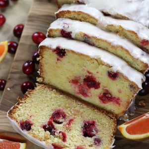 A sliced Cranberry Bread Recipe sitting on a wooden cutting board surrounded by fresh cranberries and sliced oranges.