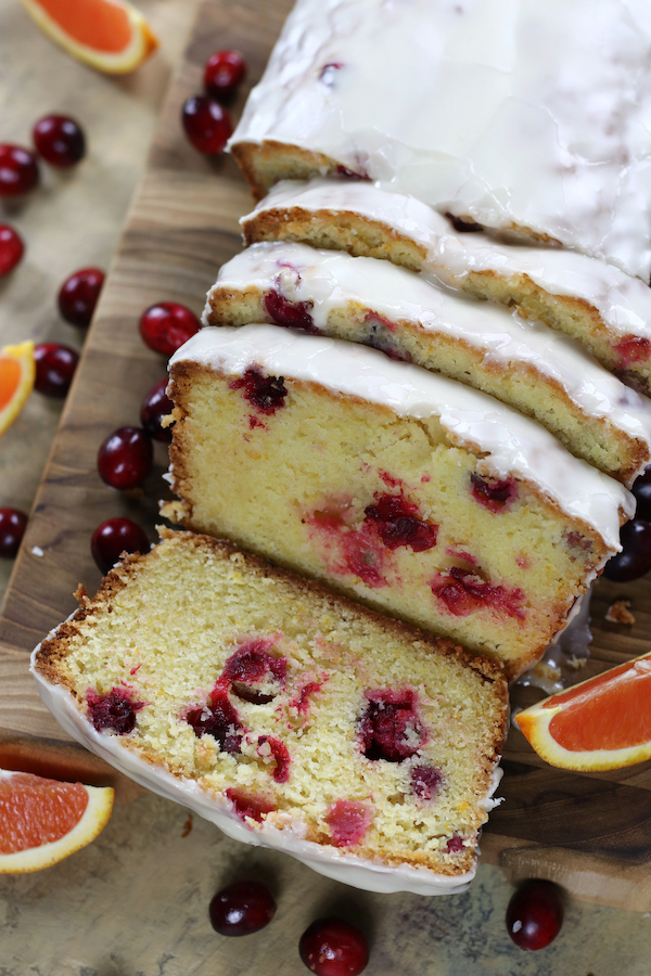 A sliced Cranberry Bread Recipe sitting on a wooden cutting board surrounded by fresh cranberries and sliced oranges.