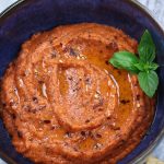 Red Pepper Dip in a blue bowl garnished with red chili pepper flakes, olive oil and basil leaves.