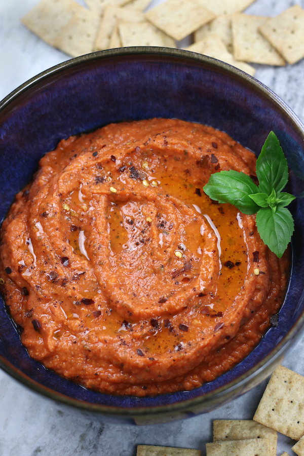 Red Pepper Dip in a blue bowl garnished with red chili pepper flakes, olive oil and basil leaves.