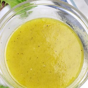 A bowl of White Balsamic Dressing on a white countertop surrounded by a few lettuce leaves.