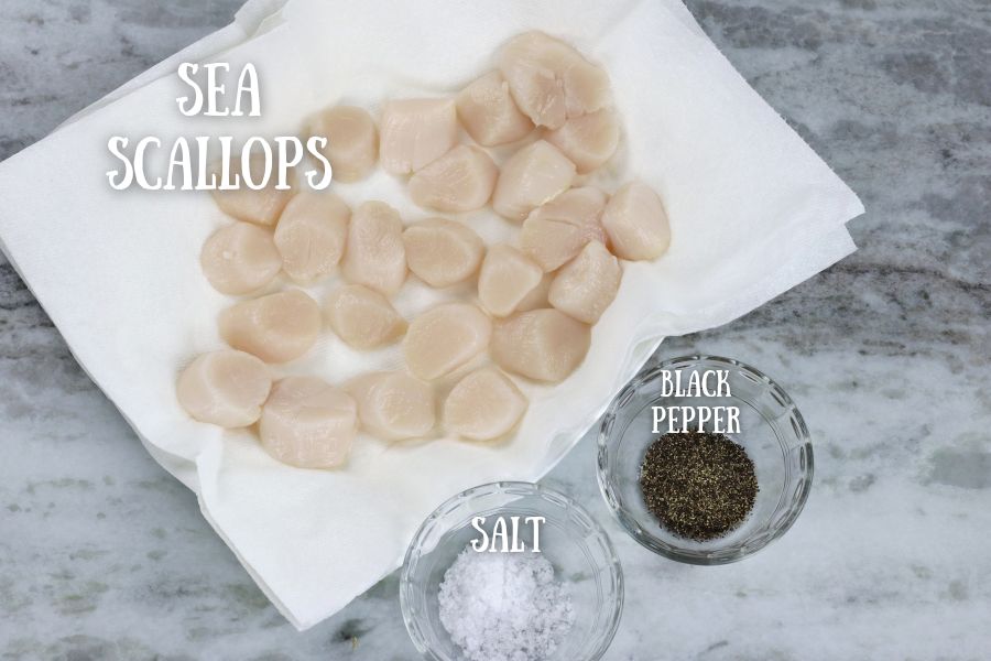 Defrosted sea scallops sitting in a single layer on a paper towel lined plate sitting alongside small bowls of salt and pepper.