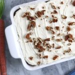 Carrot Cake with Pineapple sitting next to fresh carrots on a white marble countertop.