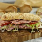 Italian sandwich filled with cured meats and crisp vegetables on a sesame roll, displayed on a wooden board with sandwiches wrapped in paper in the background.