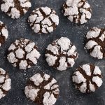 Close-up of Chocolate Crackle Cookies with crisp, snowy powdered sugar coating scattered across a dark surface.
