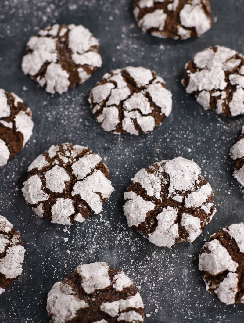 Chocolate Crinkle Cookies with crackled powdered sugar tops arranged on a dark baking sheet.