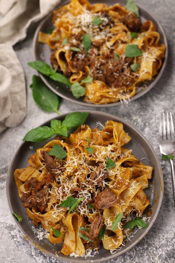Two plates full of Beef Ragu with Pappardelle pasta that are garnished with torn basil and grated parmesan.