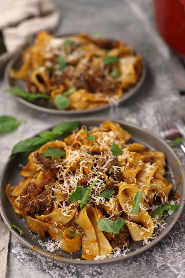 Chuck Roast Ragu recipe plated on two dishes with pappardelle and garnishes, forks alongside, with the red cooking pot partially in view.