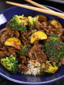 Ground Beef Teriyaki with zucchini, broccoli, mushrooms and rice served in a blue glass bowl with chopsticks on top.