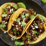 three Ground Beef Tacos on a dark grey plate surrounded by lime slices, a bowl of cut cheery tomatoes and cilantro.