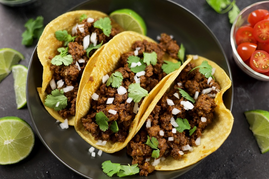 three Ground Beef Tacos on a dark grey plate surrounded by lime slices, a bowl of cut cheery tomatoes and cilantro.