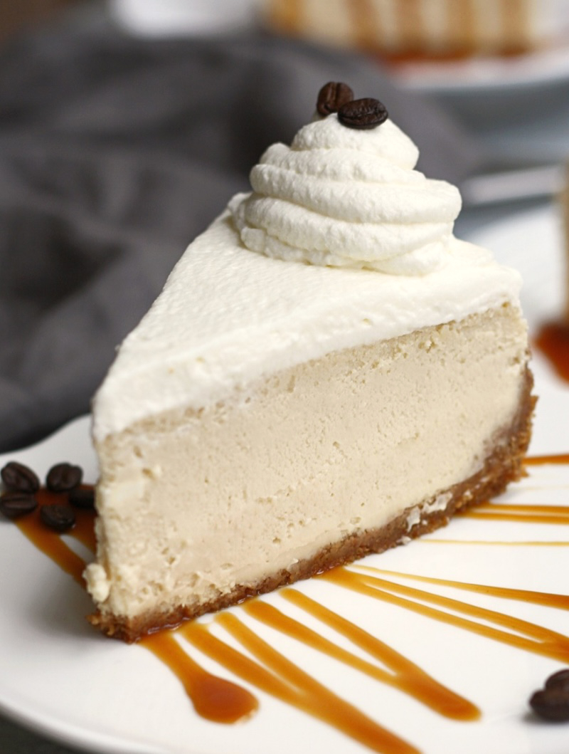 Close-up of a Caramel Macchiato Cheesecake slice topped with whipped cream and coffee beans on a caramel-drizzled plate.